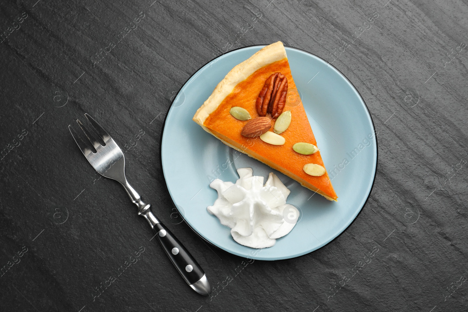 Photo of Piece of tasty homemade pumpkin pie with whipped cream, seeds, nuts and fork on black table, top view