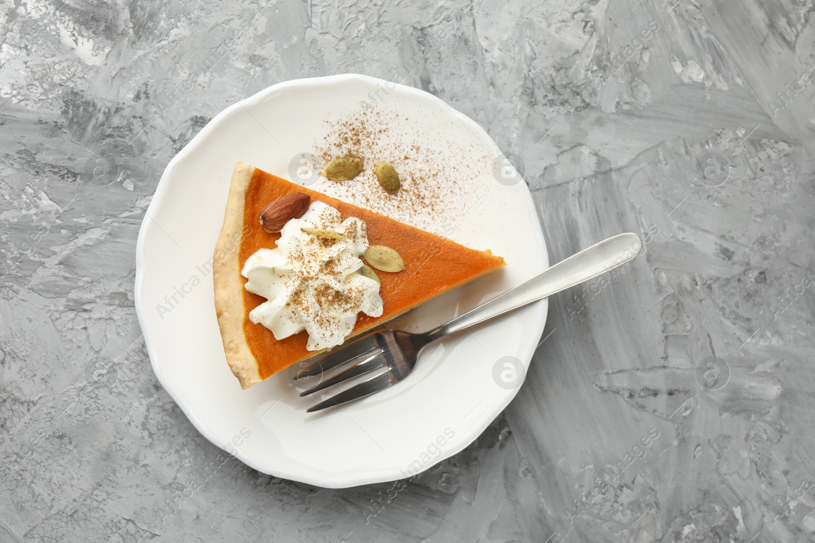 Photo of Piece of tasty homemade pumpkin pie with whipped cream, seeds and almond on grey table, top view