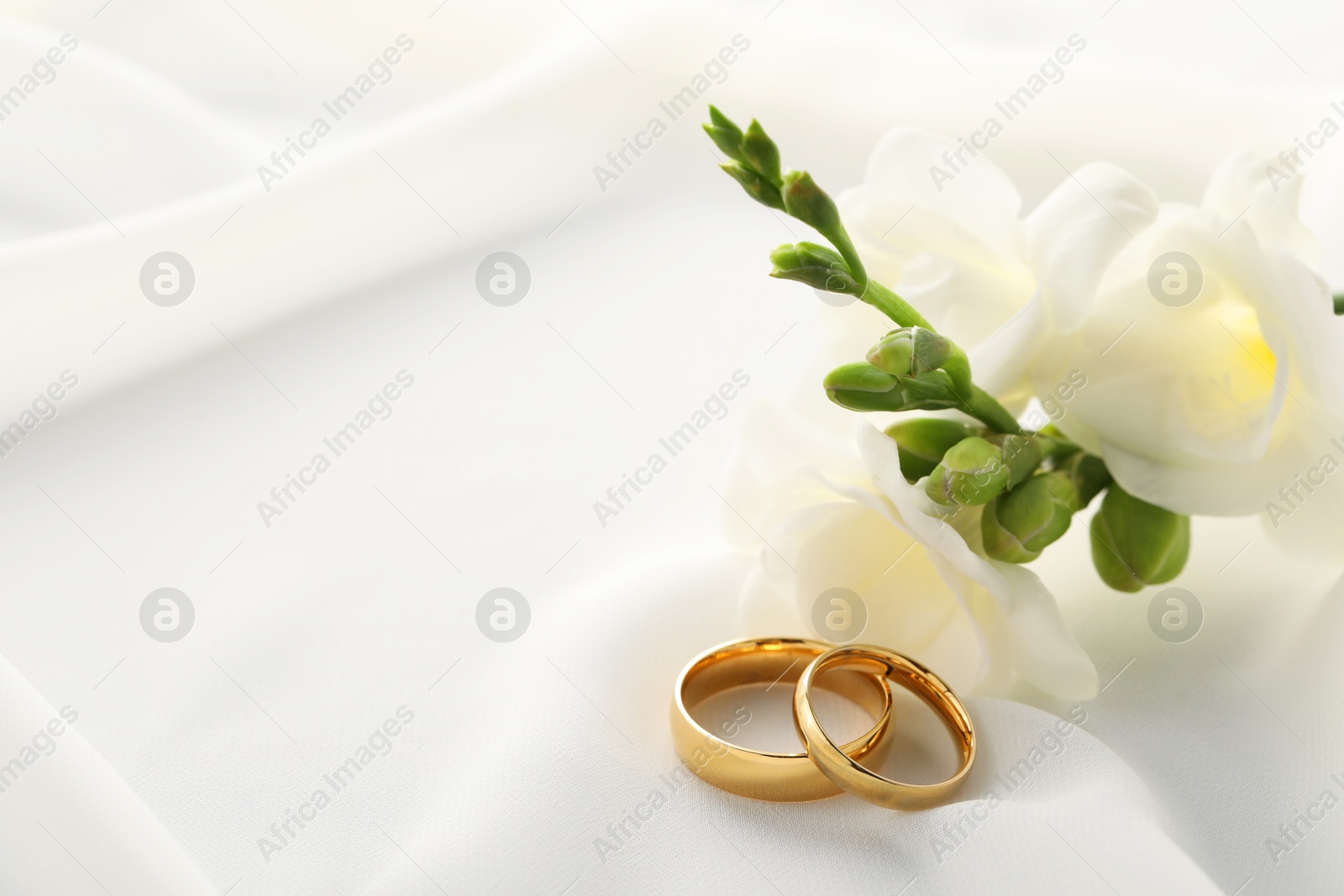 Photo of Golden wedding rings and flowers on white fabric, closeup. Space for text