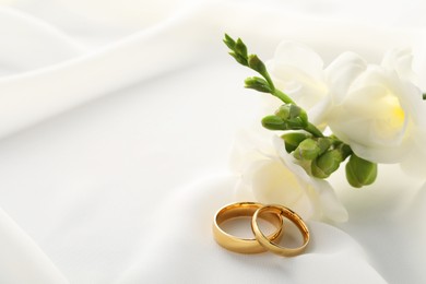 Photo of Golden wedding rings and flowers on white fabric, closeup. Space for text