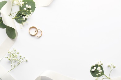 Photo of Golden wedding rings, ribbon, leaves and flowers on white background, flat lay. Space for text