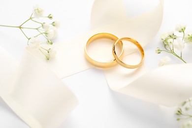 Photo of Golden wedding rings, ribbon and flowers on white background, closeup