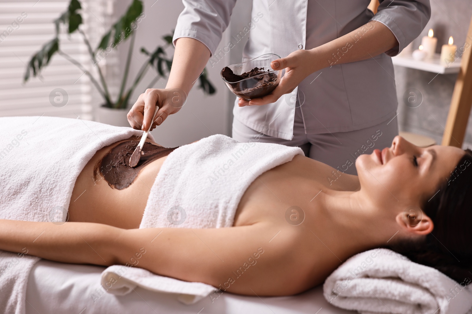 Photo of Chocolate body wrap. Spa worker applying mask onto woman's belly in salon, closeup