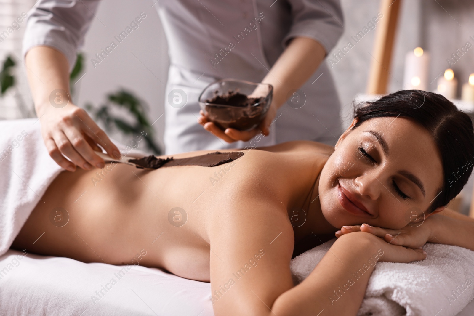 Photo of Chocolate body wrap. Spa worker applying mask onto woman's back in salon, closeup