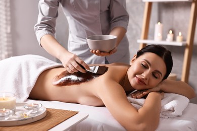 Photo of Chocolate body wrap. Spa worker applying mask onto woman's back in salon, closeup