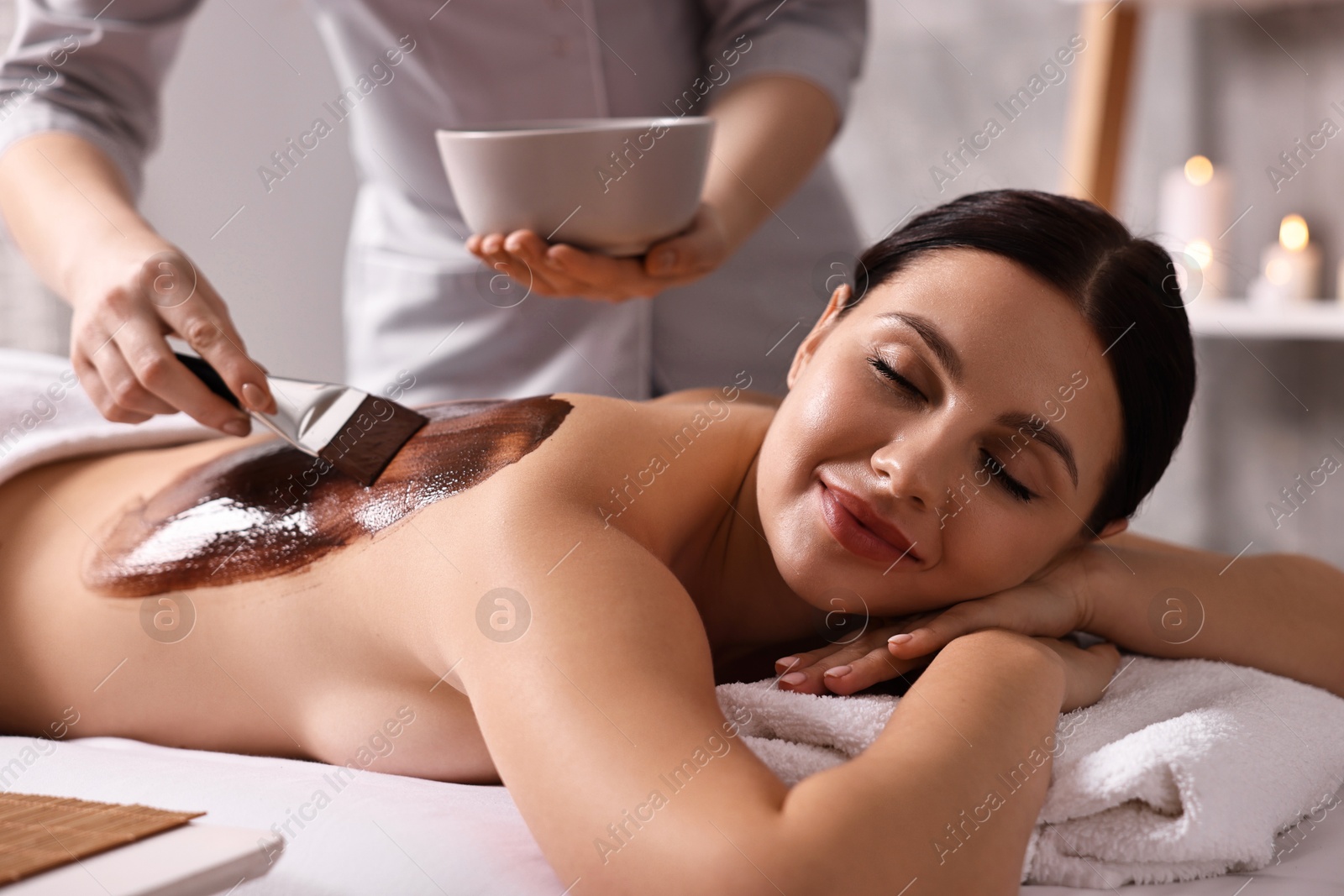 Photo of Chocolate body wrap. Spa worker applying mask onto woman's back in salon, closeup