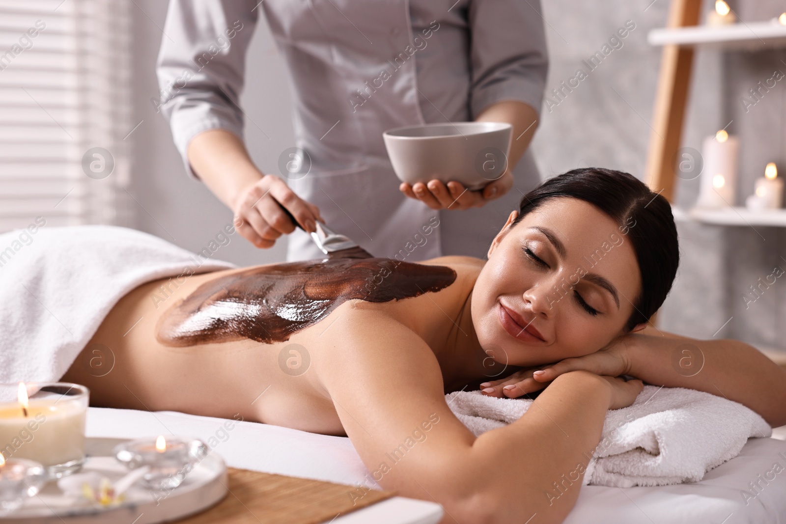 Photo of Chocolate body wrap. Spa worker applying mask onto woman's back in salon, closeup