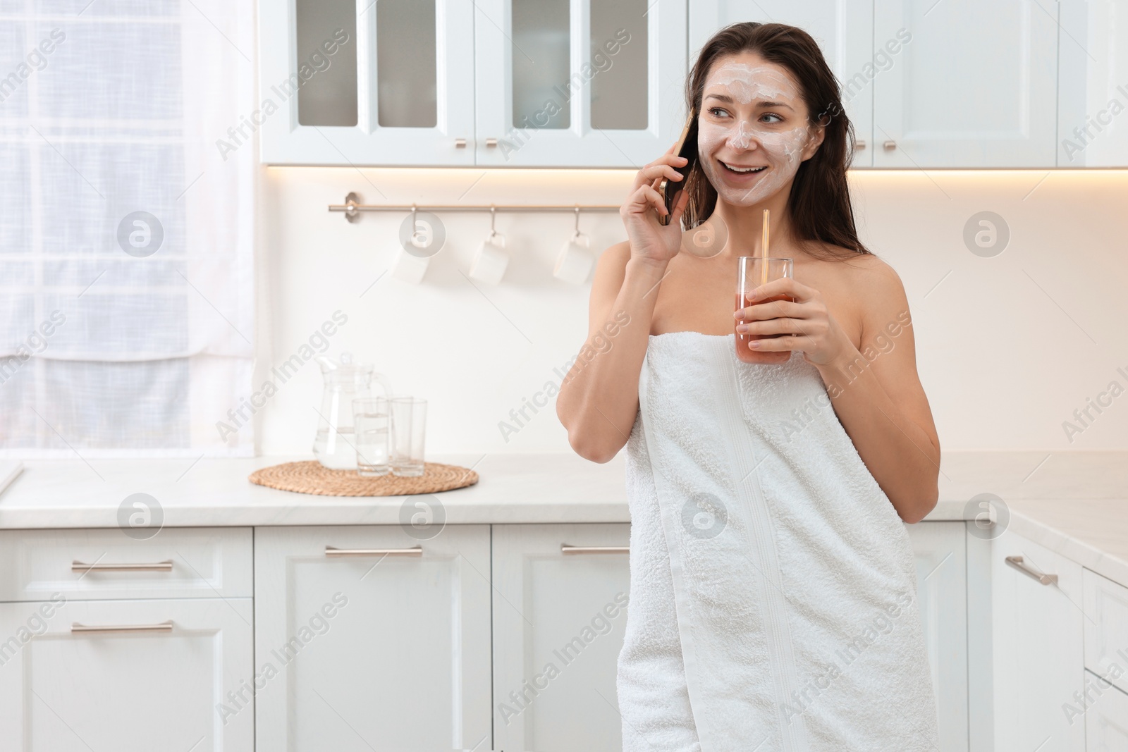 Photo of Spa day. Beautiful woman with face mask and juice talking on smartphone in kitchen, space for text