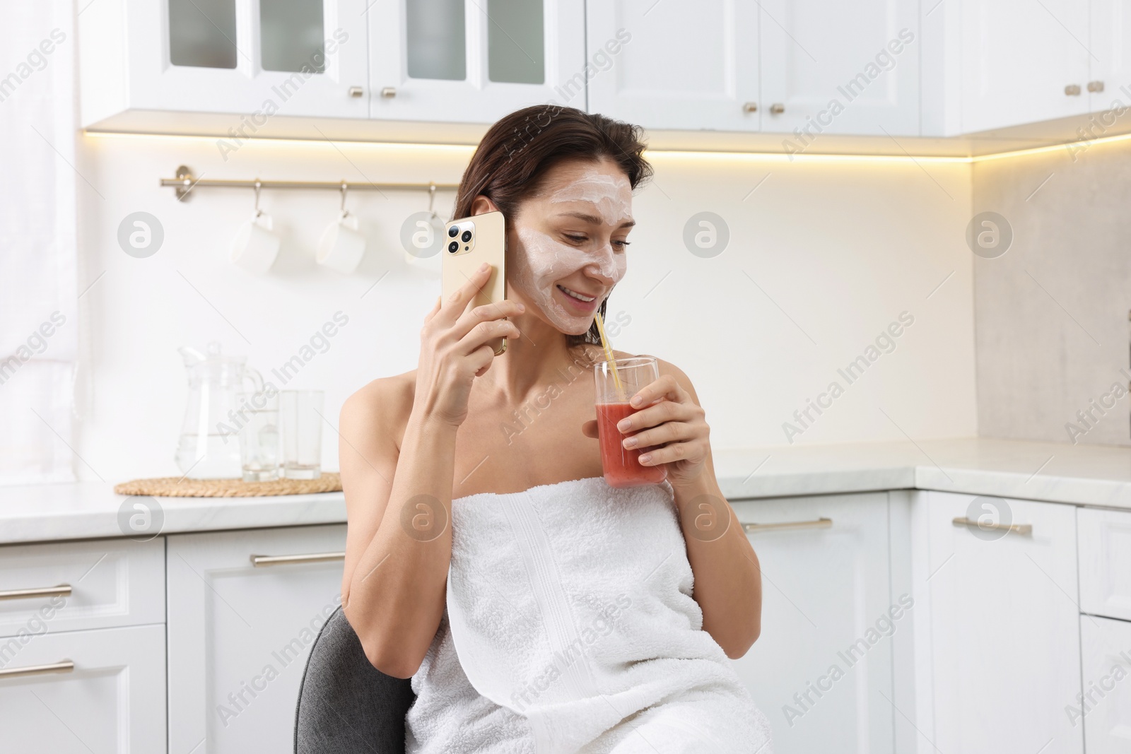 Photo of Spa day. Beautiful woman with face mask and juice talking on smartphone in kitchen