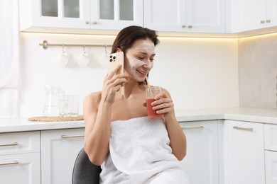 Spa day. Beautiful woman with face mask and juice talking on smartphone in kitchen