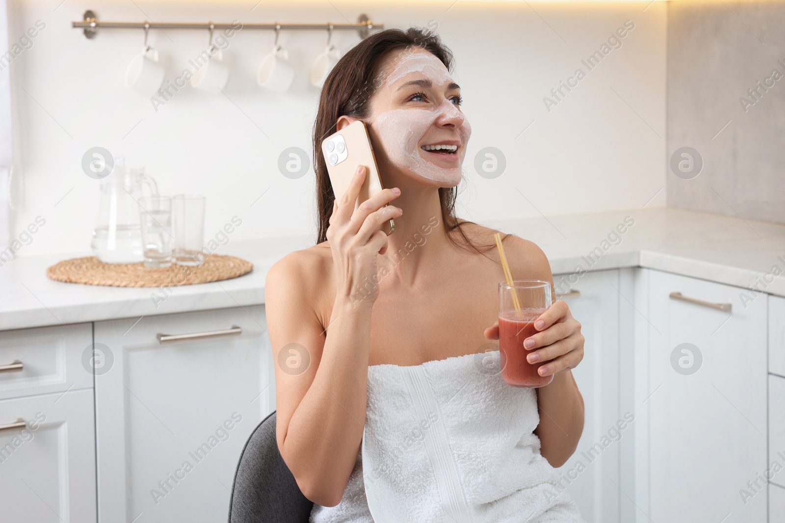 Photo of Spa day. Beautiful woman with face mask and juice talking on smartphone in kitchen