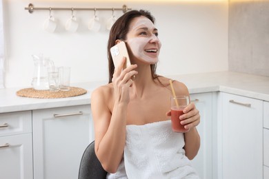 Spa day. Beautiful woman with face mask and juice talking on smartphone in kitchen