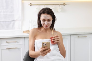 Spa day. Beautiful woman with face mask and juice using smartphone in kitchen