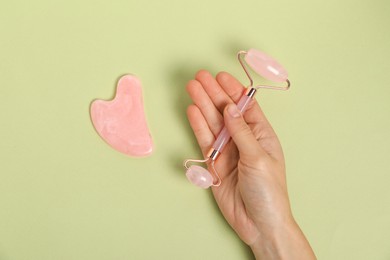 Woman with face roller and gua sha tool on light green background, top view