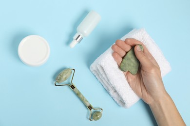 Photo of Woman with gua sha tool, face roller, cosmetic products and towel on light blue background, top view