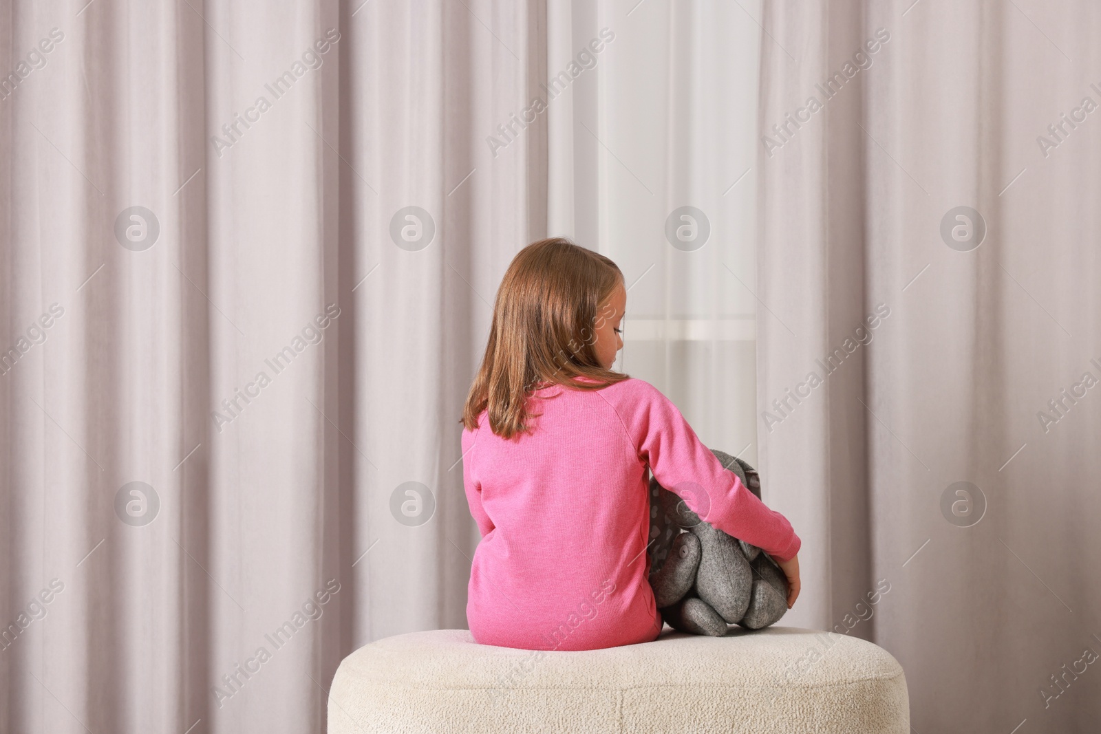 Photo of Autism concept. Lonely little girl with toy bunny on pouf at home