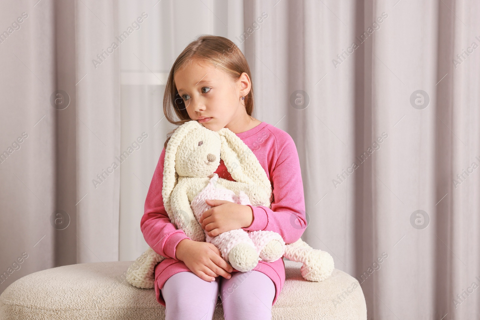 Photo of Autism concept. Lonely little girl with toy bunny on pouf at home