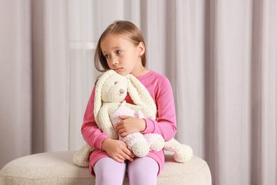 Photo of Autism concept. Lonely little girl with toy bunny on pouf at home