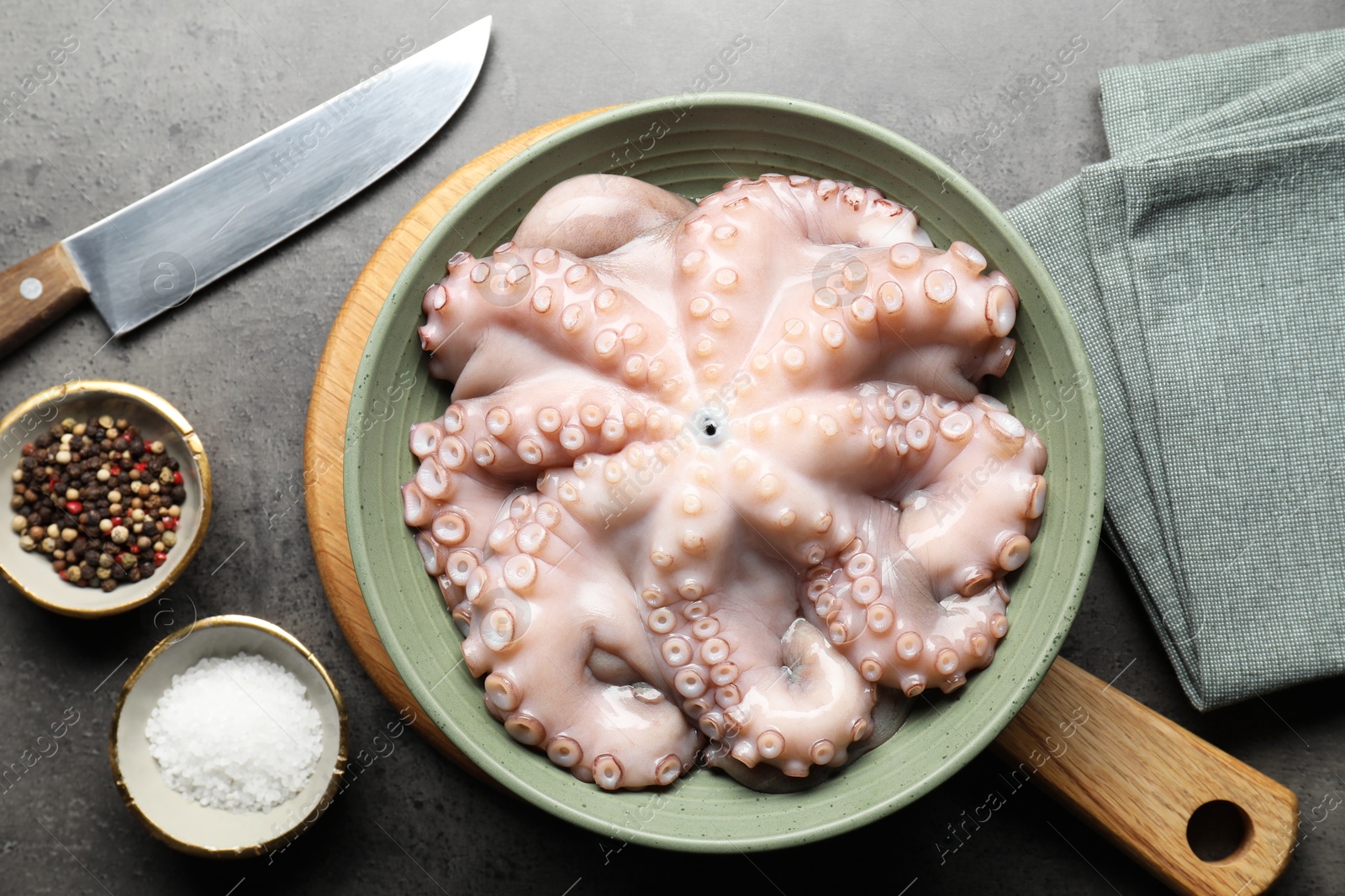 Photo of Raw octopus in bowl, peppercorns and salt on grey textured table, flat lay