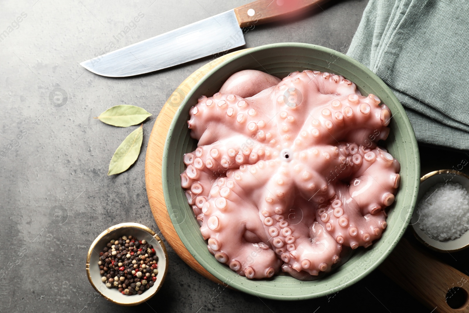 Photo of Raw octopus in bowl, spices and knife on grey textured table, flat lay