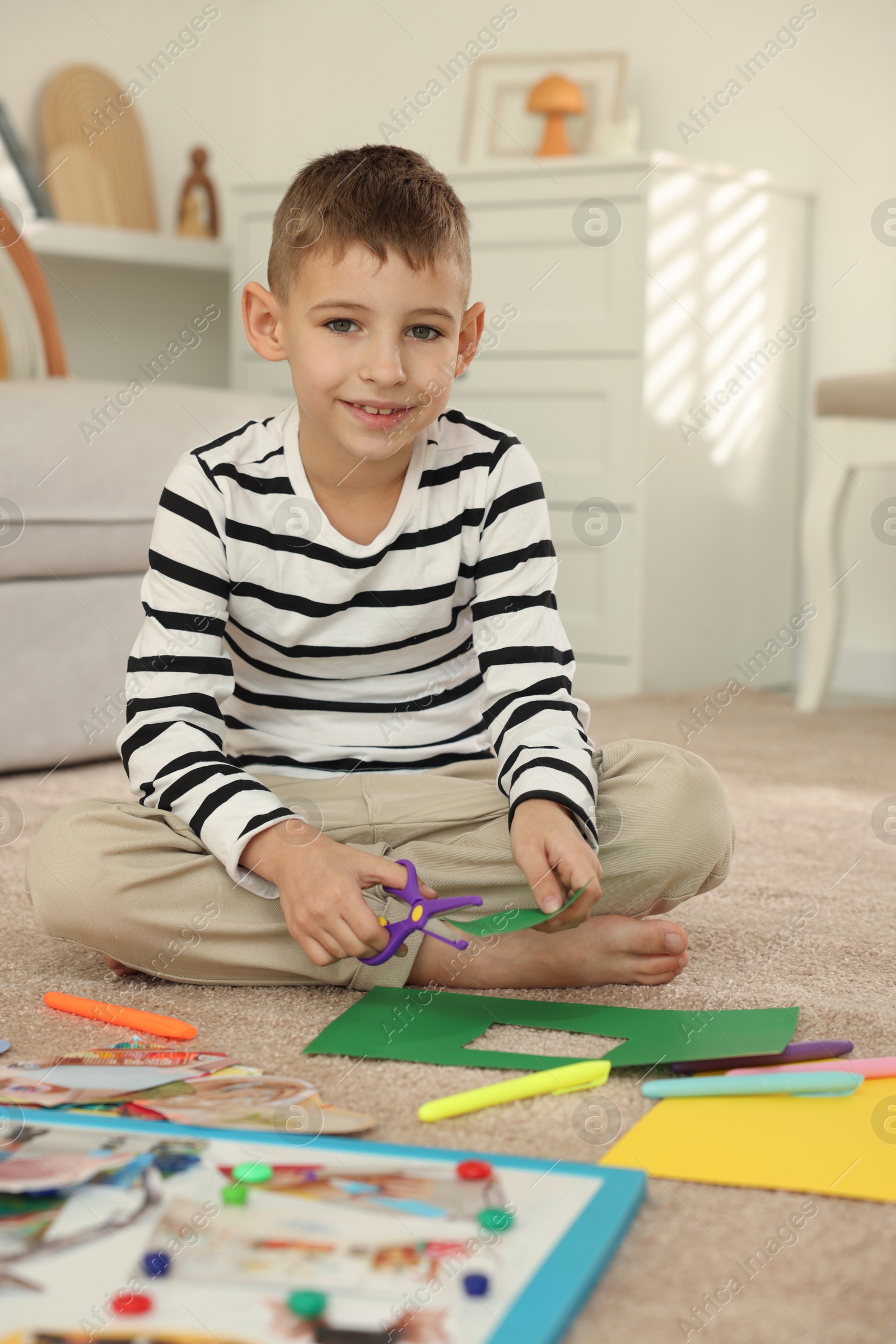 Photo of Creating vision board. Little boy cutting green paper with scissors on floor at home