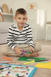 Photo of Creating vision board. Little boy cutting green paper with scissors on floor at home