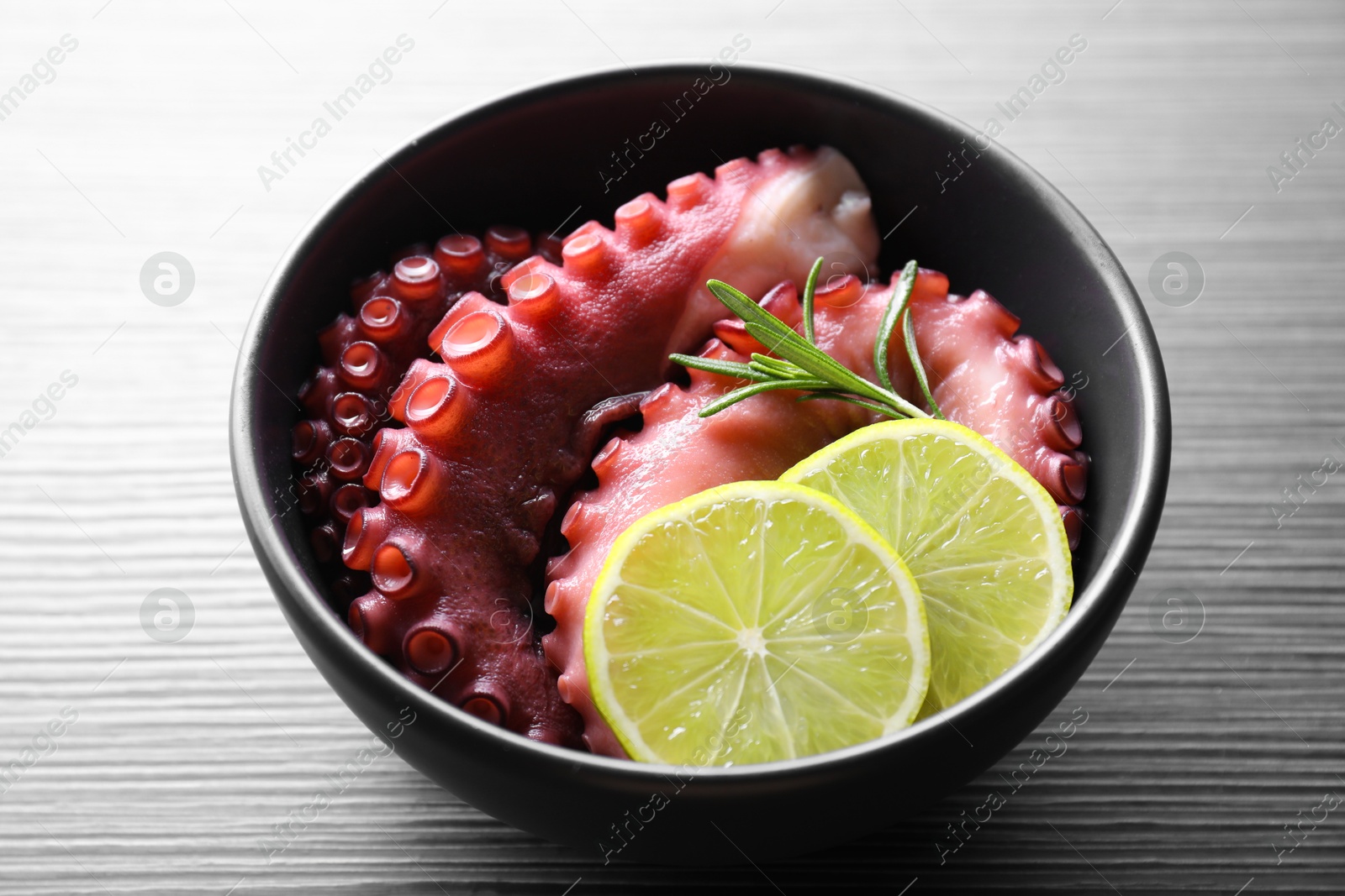 Photo of Fresh raw octopus, lemon and rosemary in bowl on dark textured table, closeup