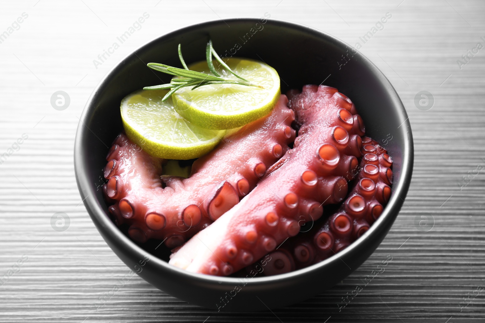 Photo of Fresh raw octopus, lemon and rosemary in bowl on dark textured table, closeup