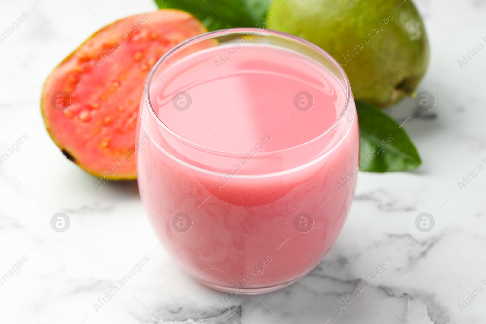 Photo of Tasty guava juice in glass, leaves and fruits on white marble table, closeup