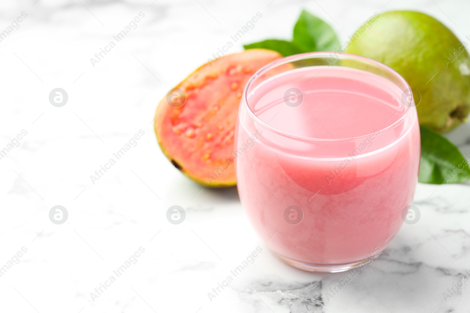 Photo of Tasty guava juice in glass, leaves and fruits on white marble table, closeup. Space for text