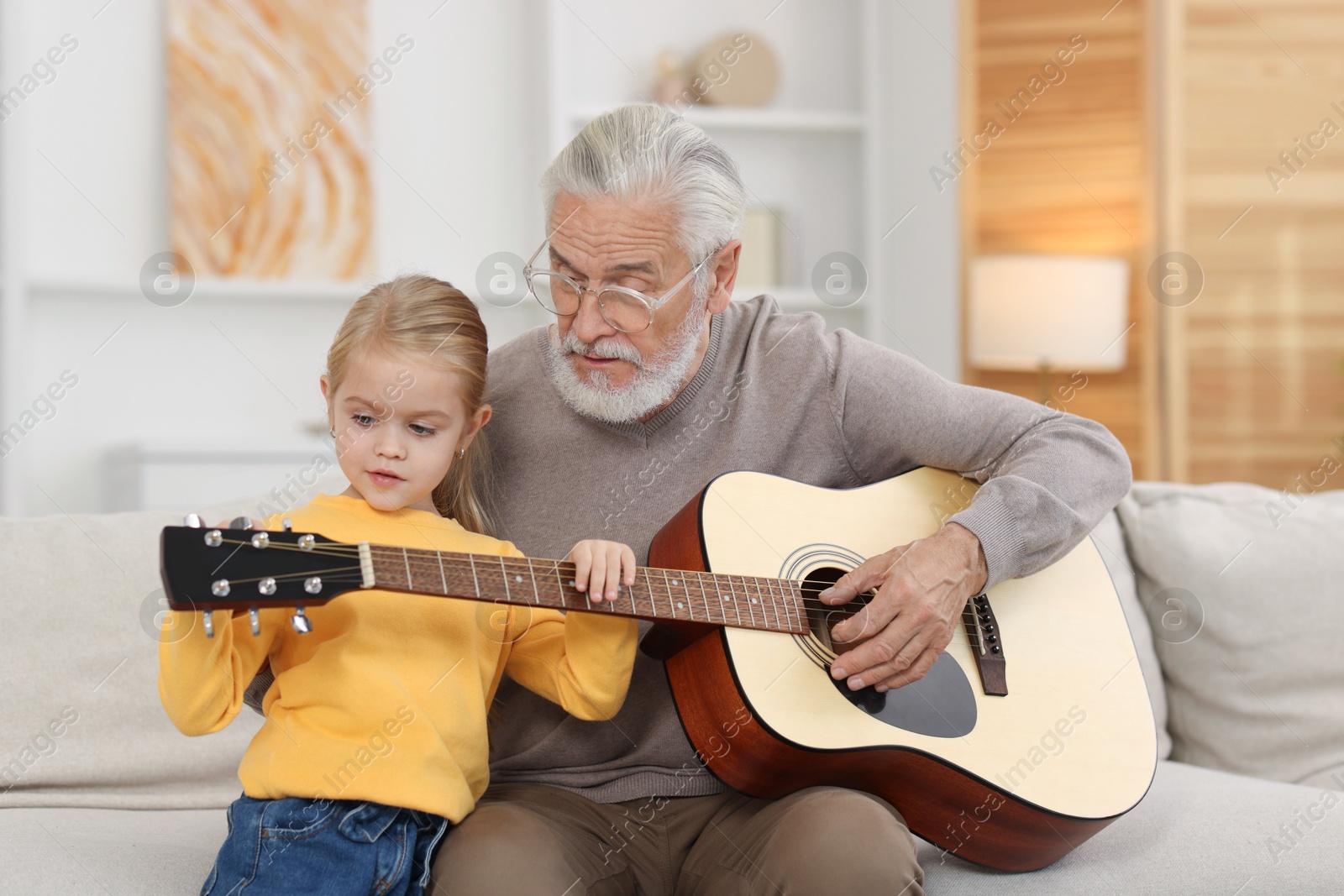 Photo of Grandpa teaching his granddaughter to play guitar on sofa at home