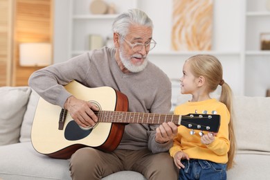 Photo of Grandpa teaching his granddaughter to play guitar on sofa at home