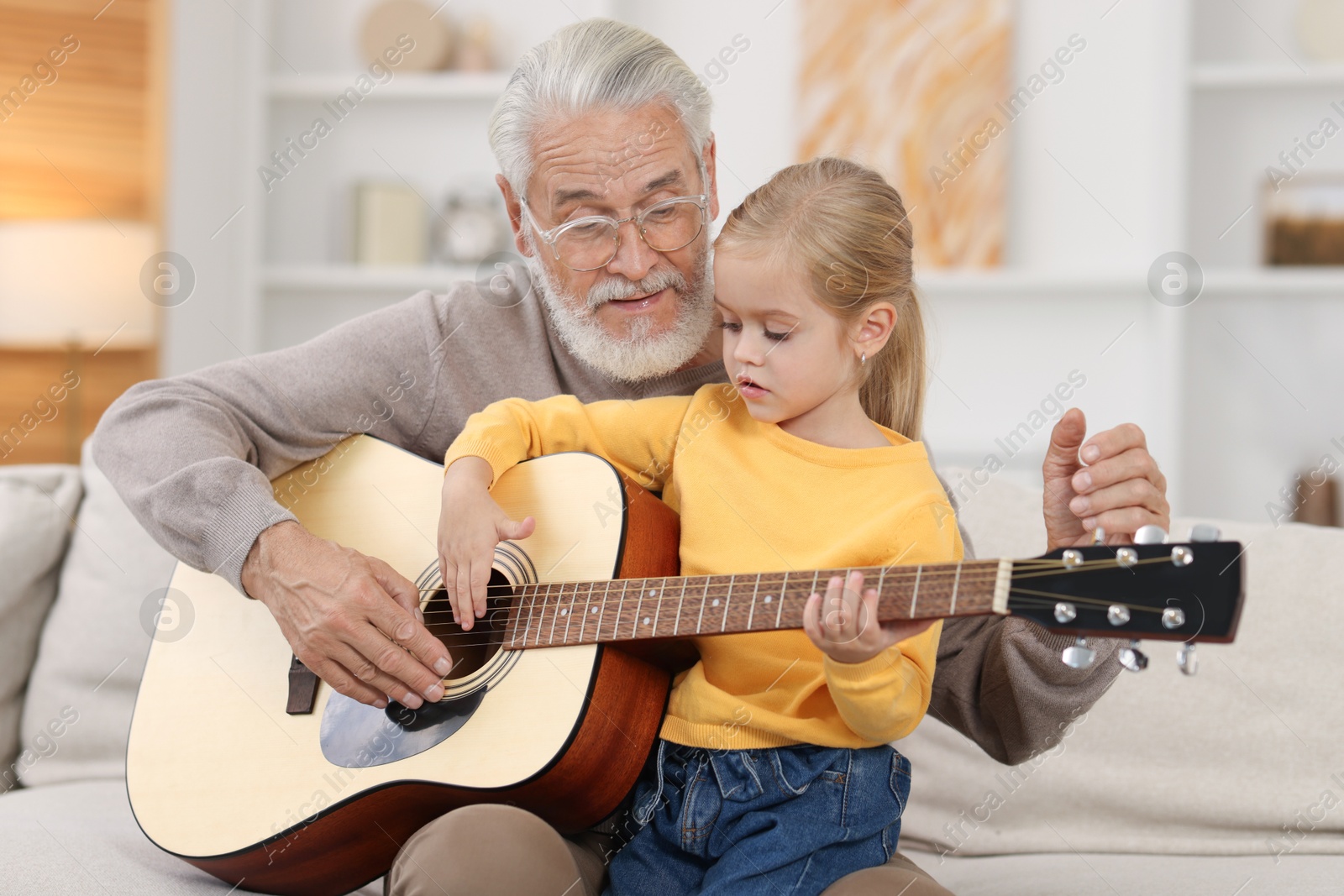 Photo of Grandpa teaching his granddaughter to play guitar on sofa at home