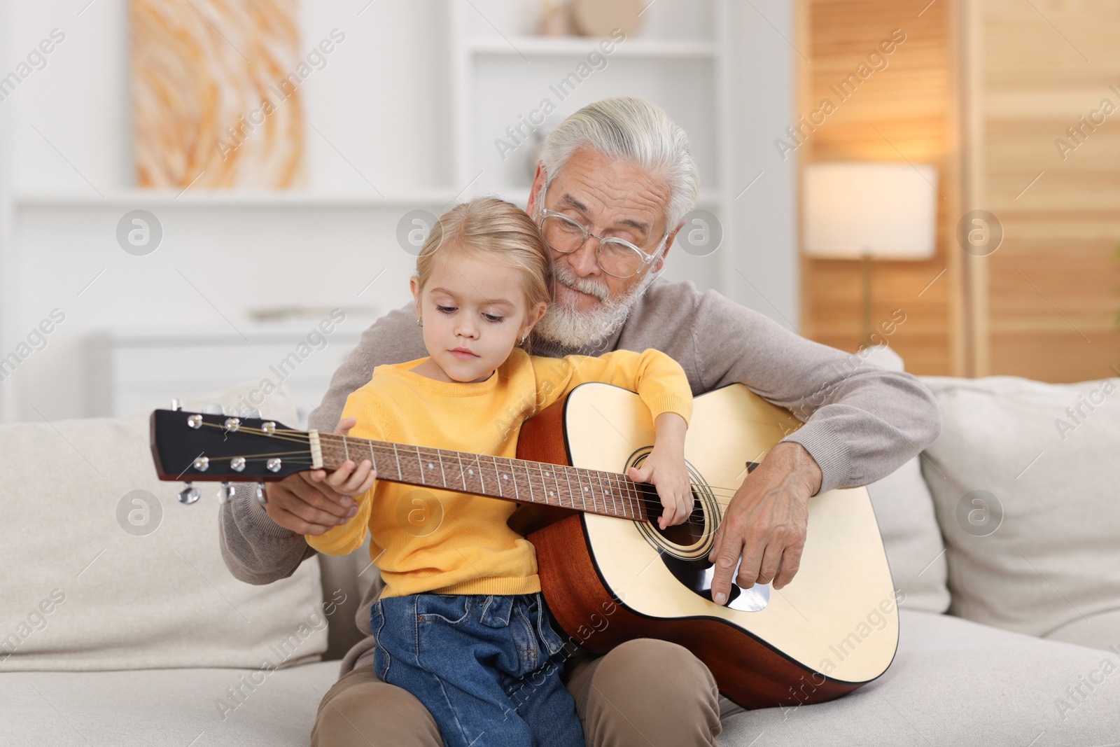 Photo of Grandpa teaching his granddaughter to play guitar on sofa at home