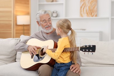 Photo of Grandpa teaching his granddaughter to play guitar on sofa at home