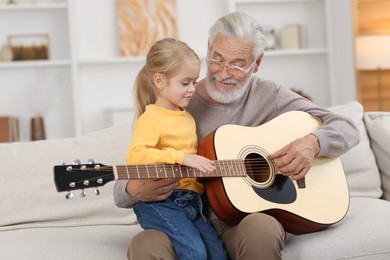 Photo of Grandpa teaching his granddaughter to play guitar on sofa at home