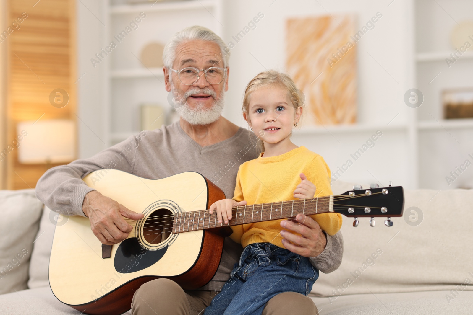 Photo of Grandpa teaching his granddaughter to play guitar on sofa at home