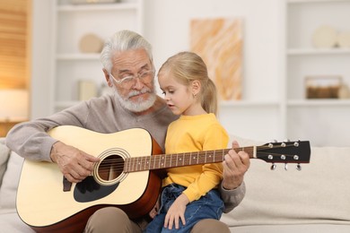 Photo of Grandpa teaching his granddaughter to play guitar on sofa at home