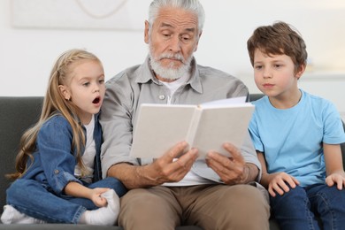 Photo of Grandpa and his grandkids reading book together on sofa at home