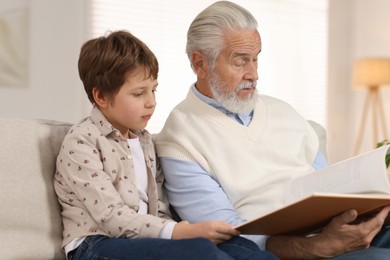 Photo of Grandpa and his grandson reading book together on sofa at home