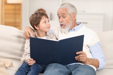 Photo of Grandpa and his grandson reading book together on sofa at home