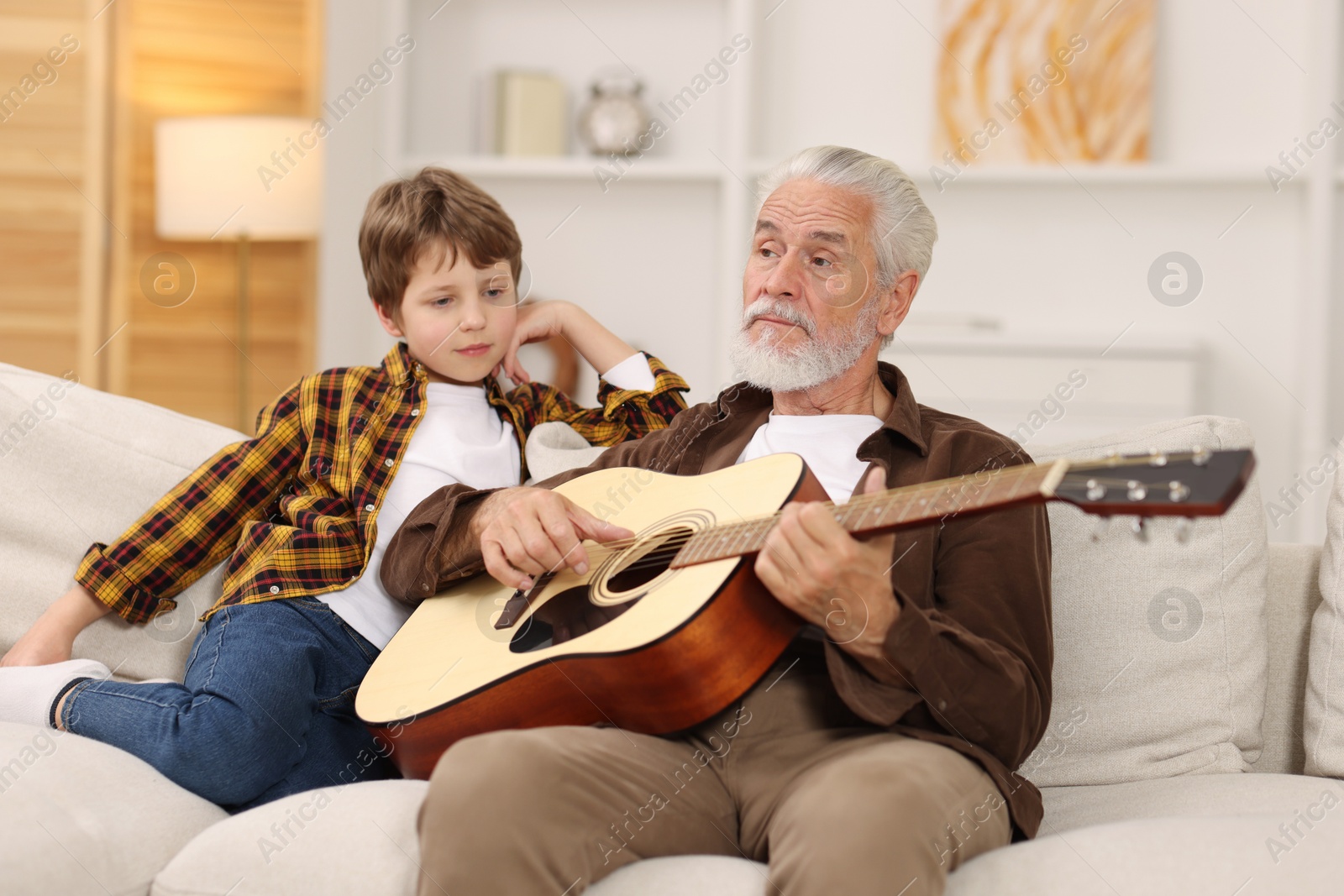 Photo of Grandpa teaching his grandson to play guitar on sofa at home