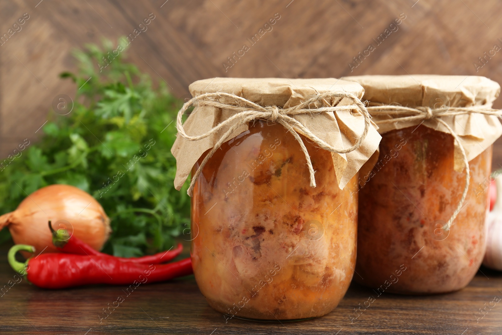 Photo of Canned meat in glass jars, chili peppers, onion and parsley on wooden table