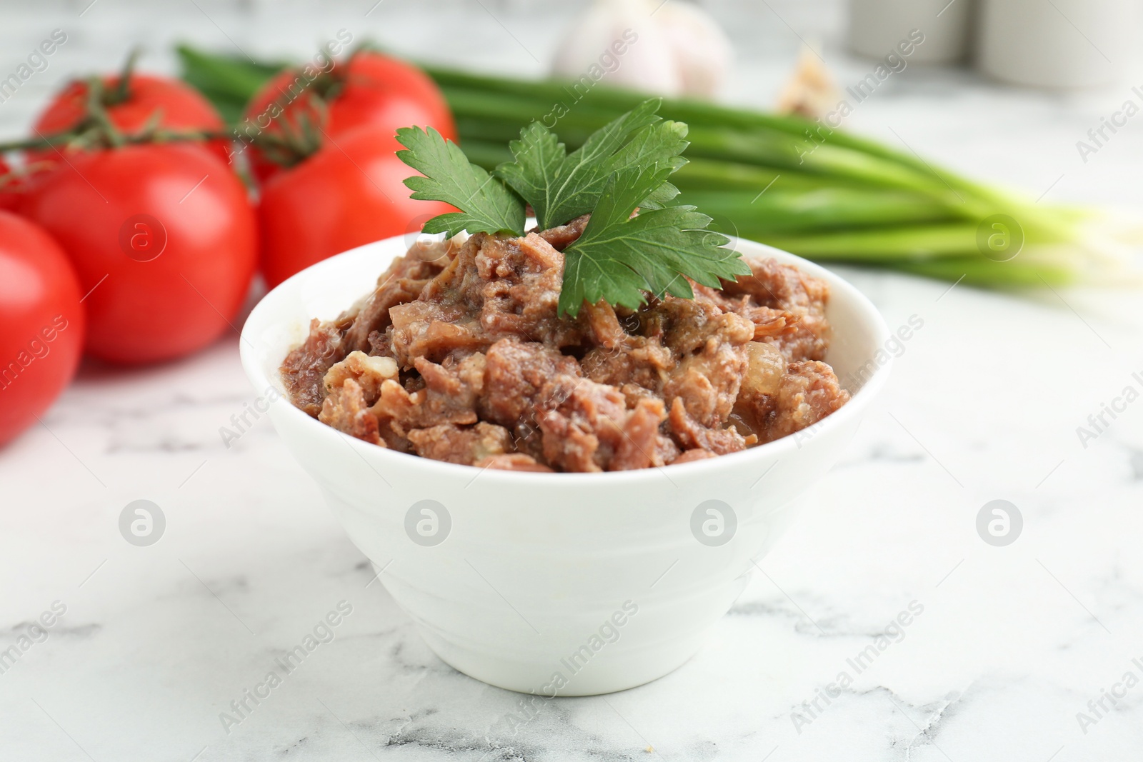 Photo of Canned meat in bowl, parsley, tomatoes and green onions on white marble table, closeup