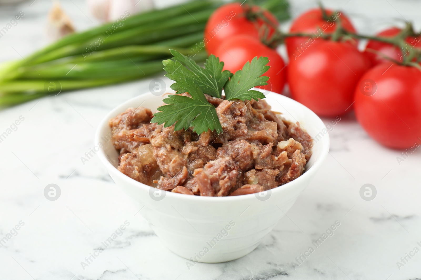 Photo of Canned meat in bowl, parsley, tomatoes and green onions on white marble table, closeup