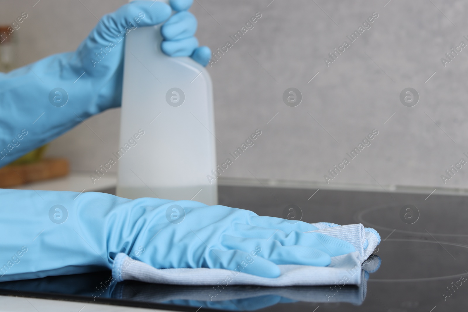 Photo of Woman using cleaning product while wiping induction cooktop with rag in kitchen, closeup