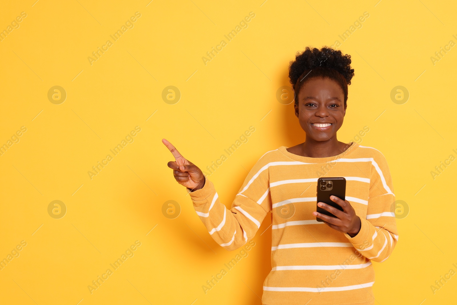 Photo of Cheerful woman with smartphone pointing at something on yellow background. Space for text