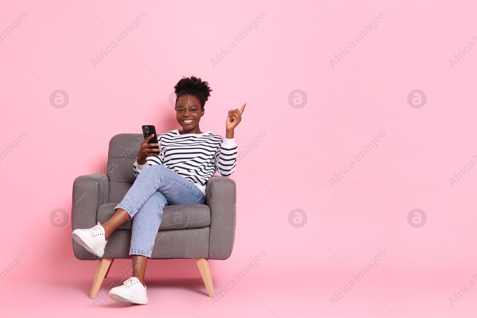 Photo of Cheerful woman with smartphone sitting in armchair and pointing at something on pink background. Space for text