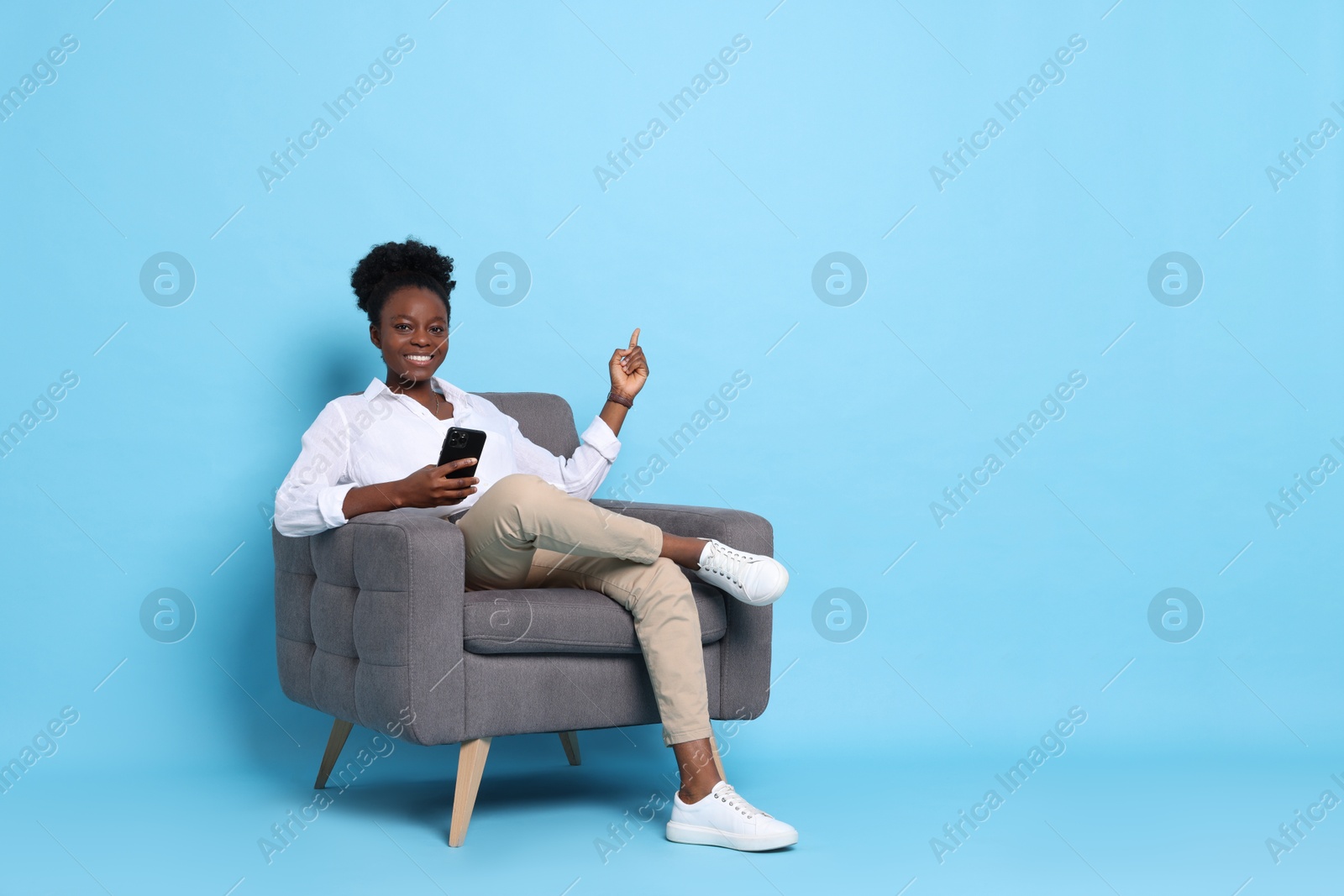 Photo of Smiling woman with smartphone sitting in armchair on light blue background. Space for text