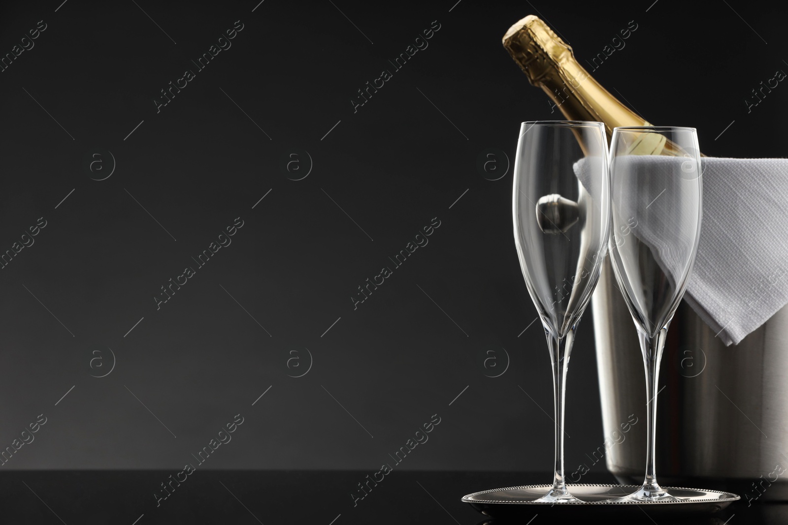 Photo of Bottle of sparkling wine in cooler bucket and glasses on black table against grey background. Space for text